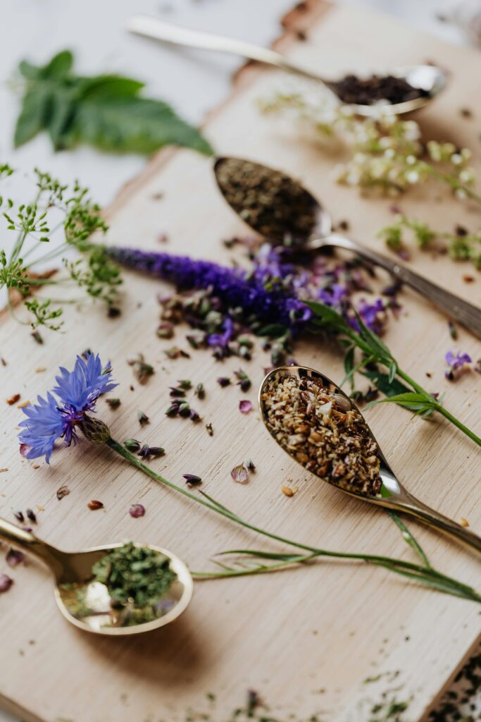 Variety of herbs and spices on a wooden board with vibrant flowers and spoons.